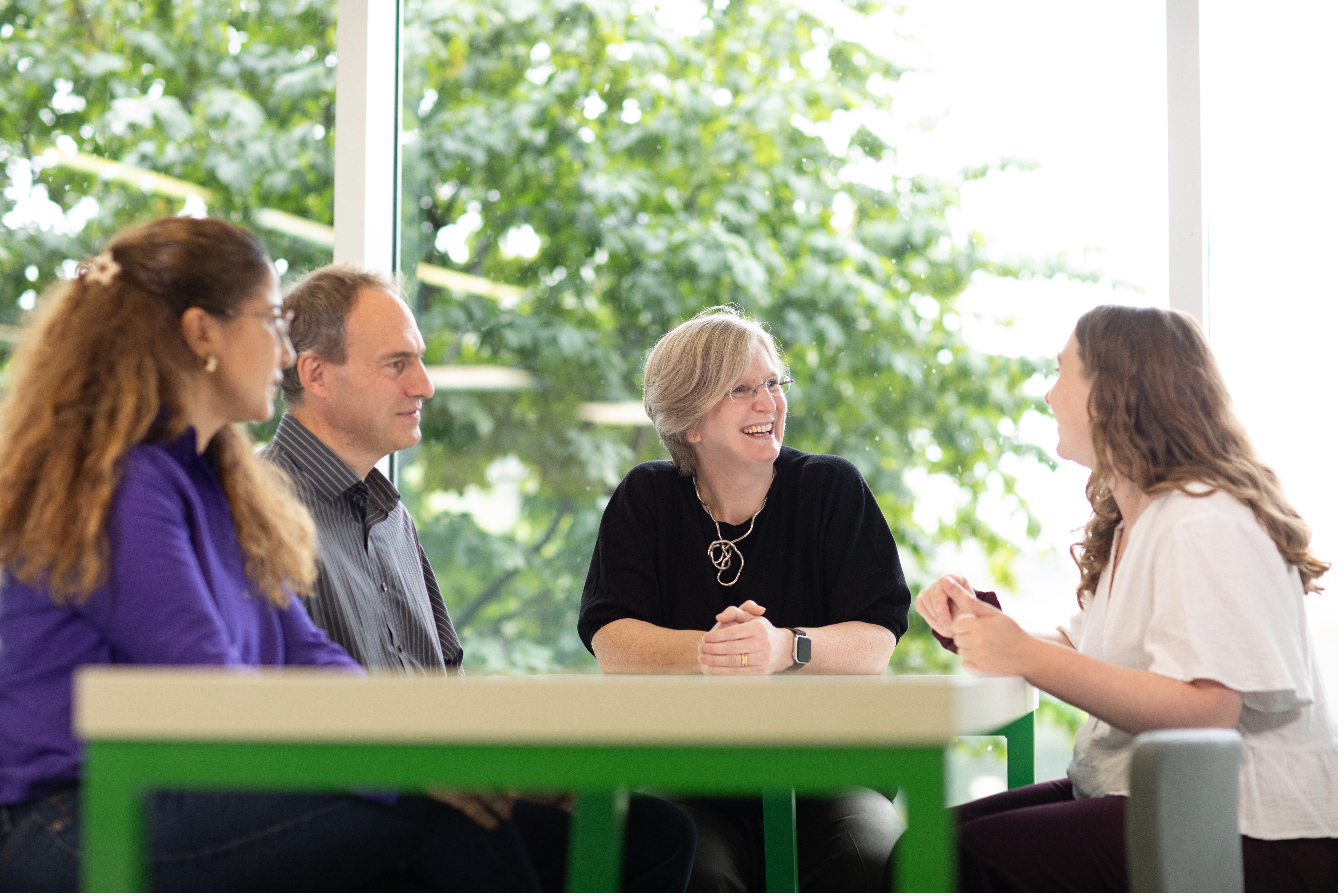 a group of people discussing and smiling around a table with a green leafy tree in the backgroundsomething